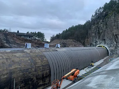 Tunnelkonstruksjon dekket av svart duk, omgitt av fjell, maskiner og byggeutstyr.