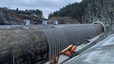 Tunnelkonstruksjon dekket av svart duk, omgitt av fjell, maskiner og byggeutstyr.